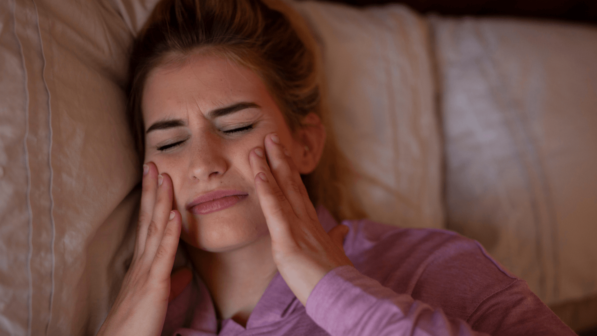 Mujer acostada en la cama tocándose las mejillas con gesto de dolor al despertar, representando la tensión mandibular causada por el bruxismo nocturno.